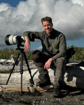 Man sitting on a log with a large camera lens on a tripod against a cloudy sky.